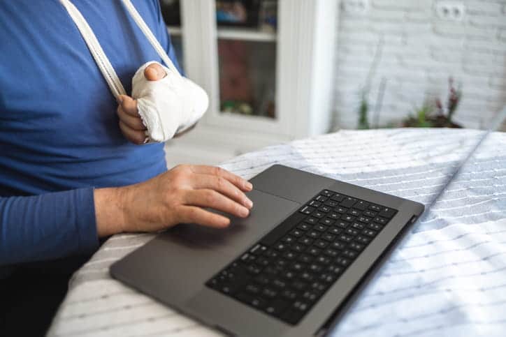 A person with their arm in a cast with a sling around their shoulder uses their laptop to contact a personal injury attorney.