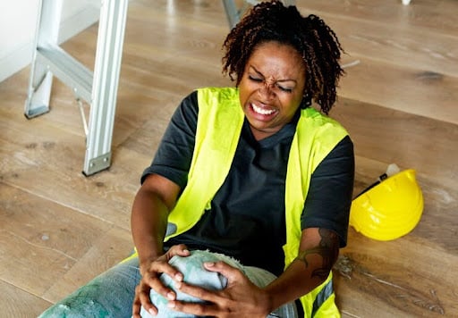 A Female Construction Worker, Wincing In Pain, Sits On The Floor Clutching Her Injured Knee.